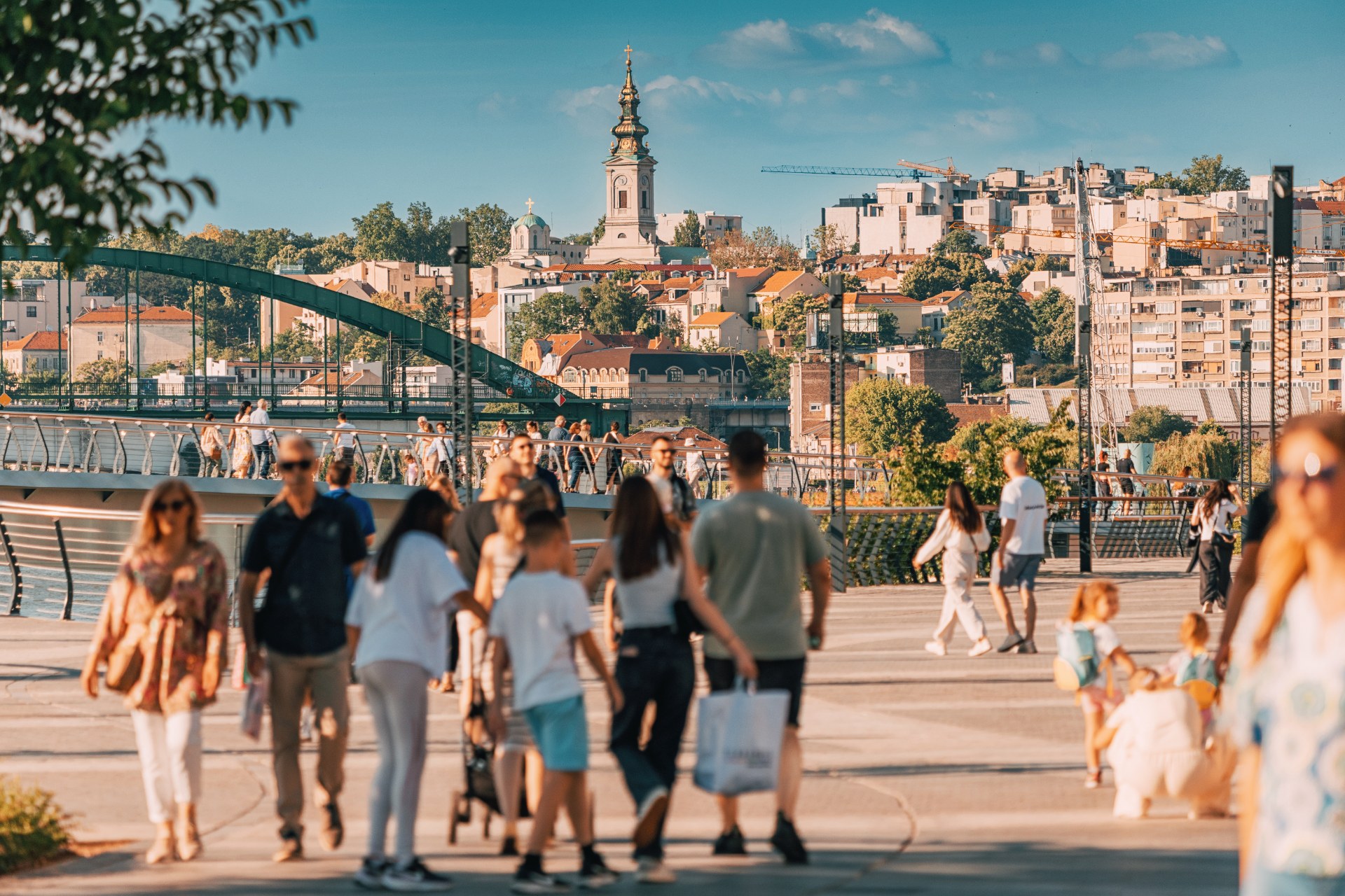 People walking on the Sava promenade in Belgrade with the old town and a church tower behind